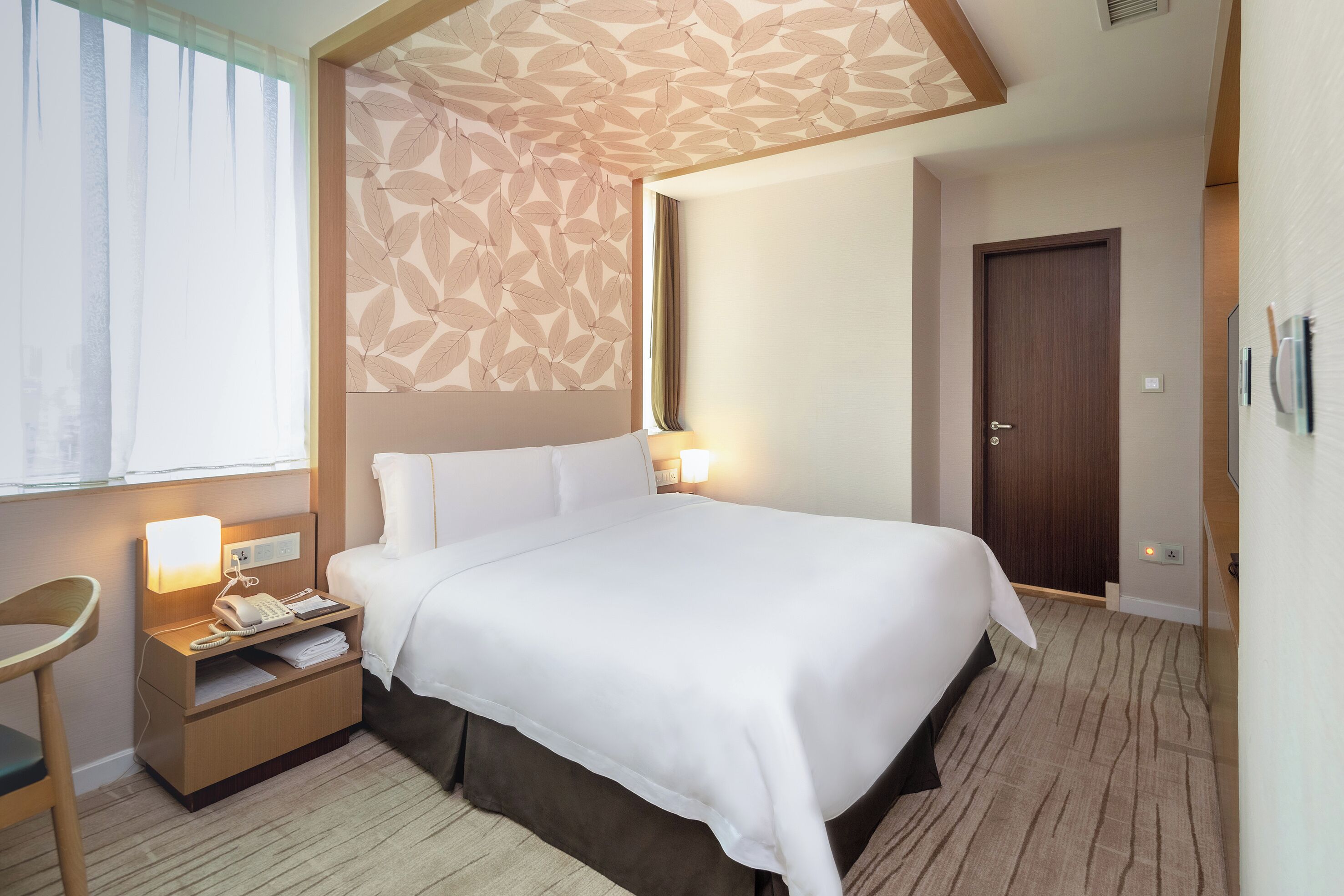 Une chambre d’hôtel avec un lit, une table de chevet avec une lampe, une chaise, une porte et un plafond décoré de motifs.