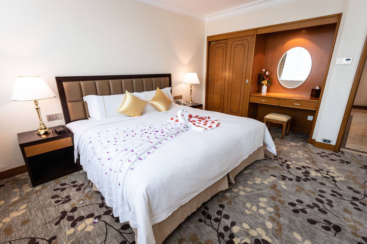 Una habitación de hotel con una cama grande, dos mesitas de noche con lámparas, un armario de madera, un pequeño banco y un espejo.