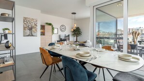 A modern dining area with a long table set for six, surrounded by chairs, a clock, and a city view through large windows. - Parkville with parking & terrace by Interhome (Blankenberge)
