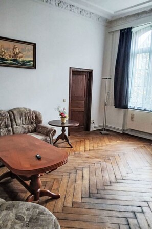 Ein Zimmer mit Fischgrätparkett, einer Couch, einem Holztisch, einem kleinen runden Tisch mit Blumenvase, einem Gemälde an der Wand, einer Tür und einer Stehlampe.
