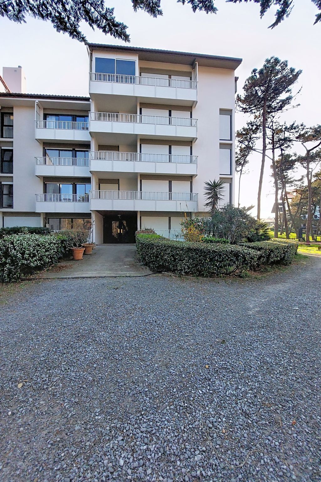Un immeuble à plusieurs étages doté de balcons, une allée gravillonnée et une végétation luxuriante tout autour.