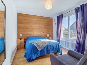 Ein Schlafzimmer mit einem hölzernen Kopfende, einem Bett mit blauer Bettwäsche, einem Nachttisch, einer Lampe, einem Fenster mit Vorhängen und einem Sessel.