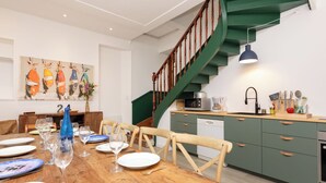 Een eetgedeelte met een houten tafel gedekt voor een maaltijd, een keuken met groene kasten en een houten trap, en een schilderij aan de muur.