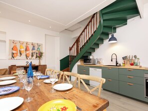 Ein Essbereich mit einem Holztisch, gedeckt für eine Mahlzeit, eine Küche mit grünen Schränken und eine Holztreppe sowie ein Gemälde an der Wand.