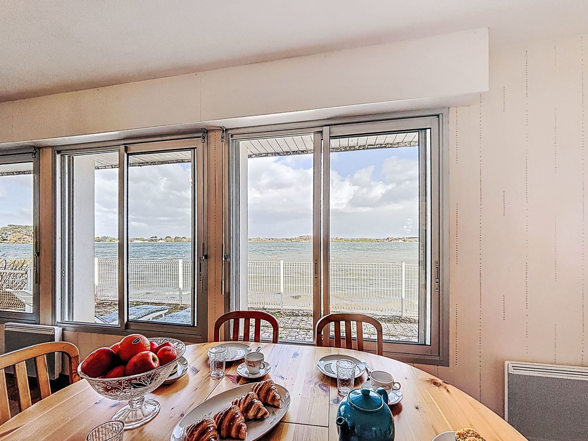 Una mesa de comedor con alimentos para el desayuno, vista al mar a través de amplias ventanas y una silla.