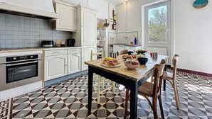 Una cocina con armarios blancos, piso de baldosas y una zona de comedor con una mesa para dos.