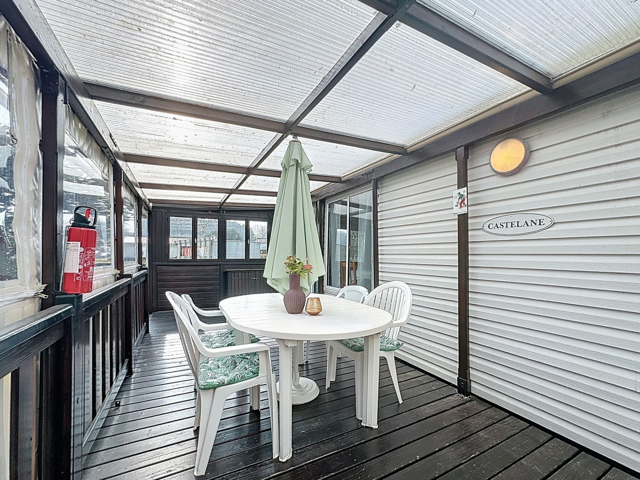Eine überdachte Terrasse im Freien mit Tisch und Stühlen, einem Feuerlöscher und einem 'CASTELANE'-Schild an der Wand.