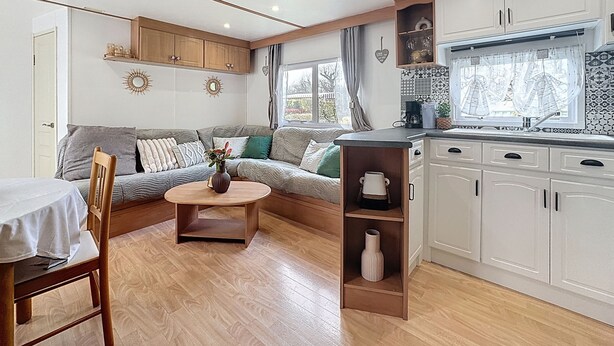 Een gezellig, compact ingerichte ruimte met een bank, een eettafel, een keuken met witte kasten en een raam voorzien van gordijnen.