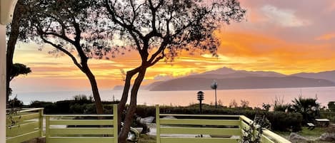 A view through a covered area with a yellow railing, overlooking a sunset over a body of water with mountains in the distance.