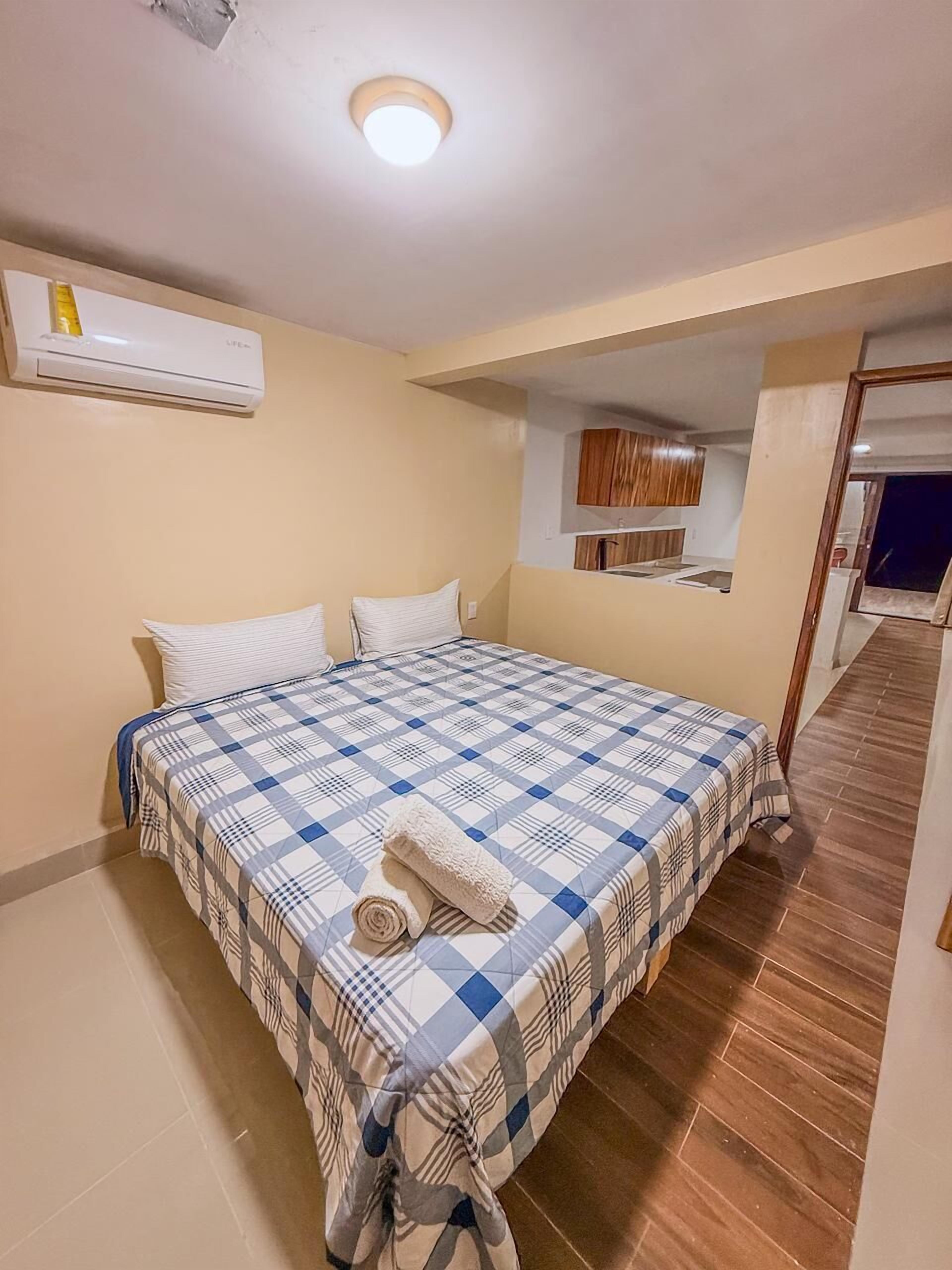 A bedroom with a bed, pillows, a rolled towel, an air conditioning unit, and a wooden floor.
