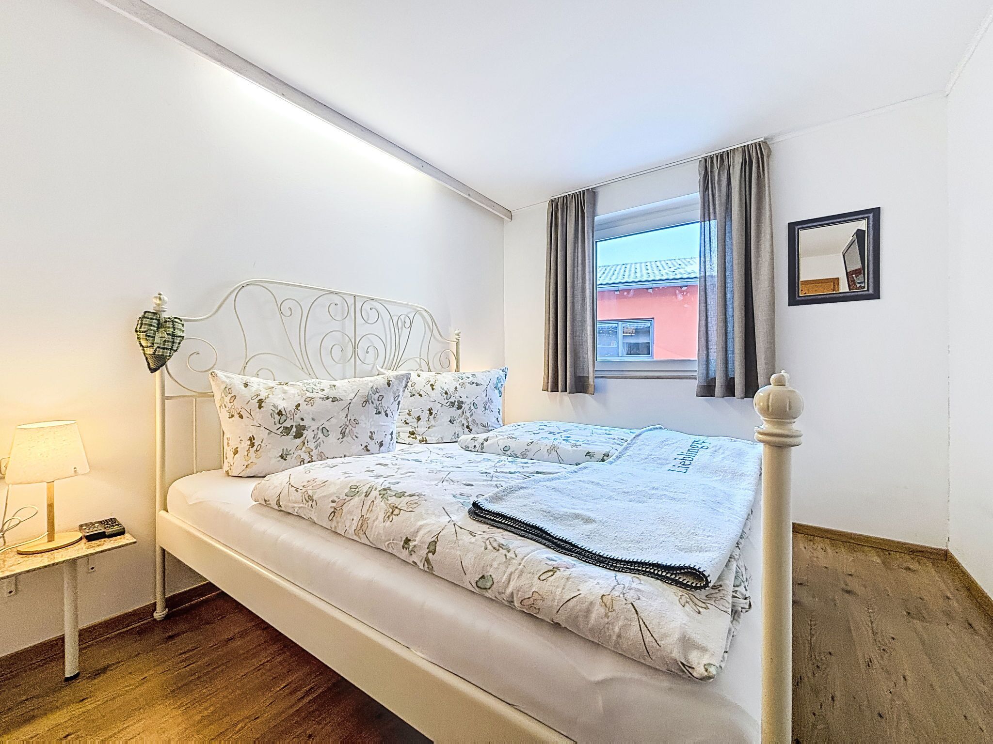 Een net opgemaakt bed met bloemenbedrukking, een bedlampje, een klein tafeltje met een afstandsbediening en een raam met gordijnen.