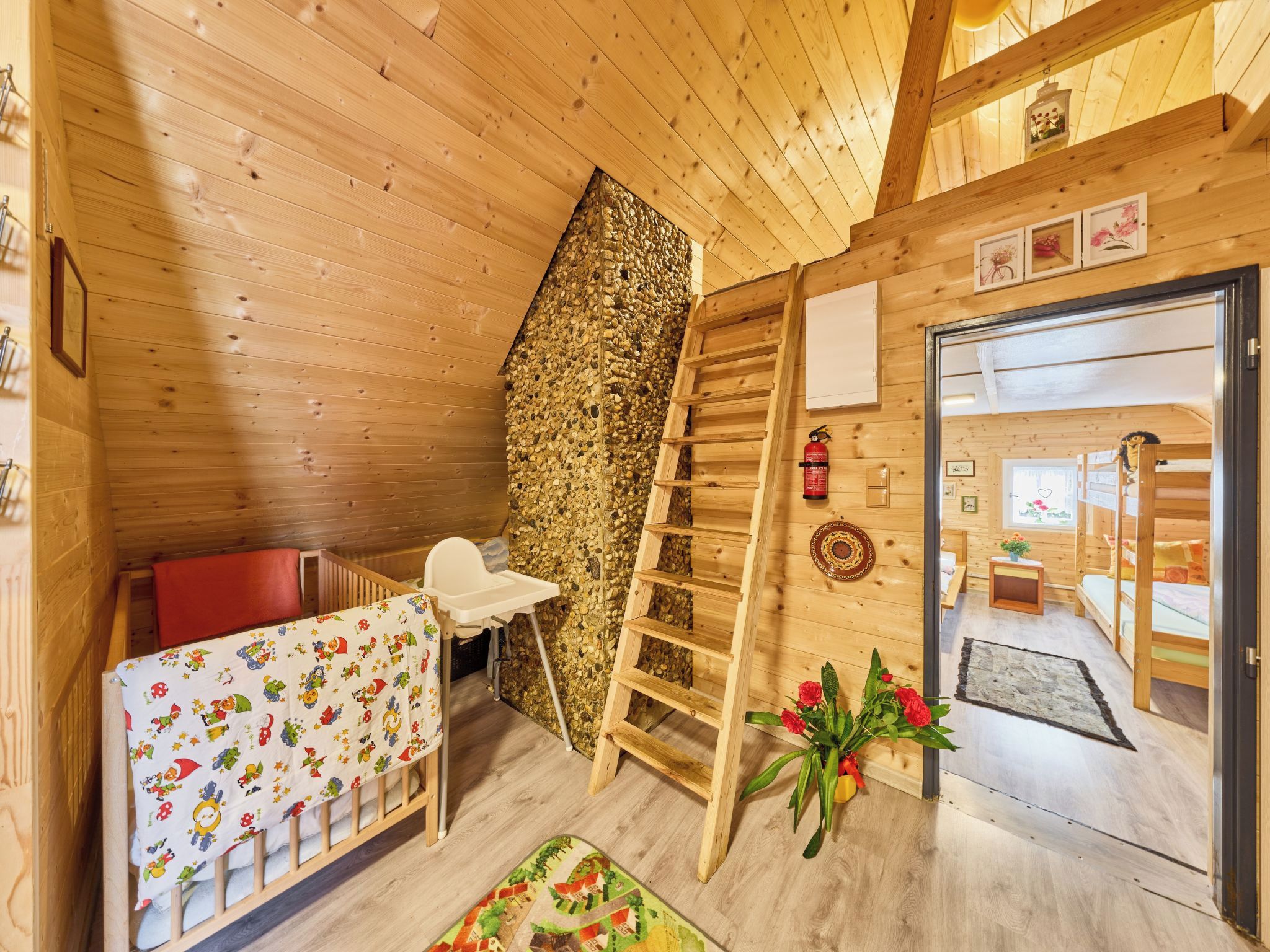 Ein gemütliches Blockhaushaus-Interieur mit Holzbalkendecke, Steinwand, einer Holzleiter, einem Babybett, einem Stuhl und einer Tür zu einem anderen Raum.