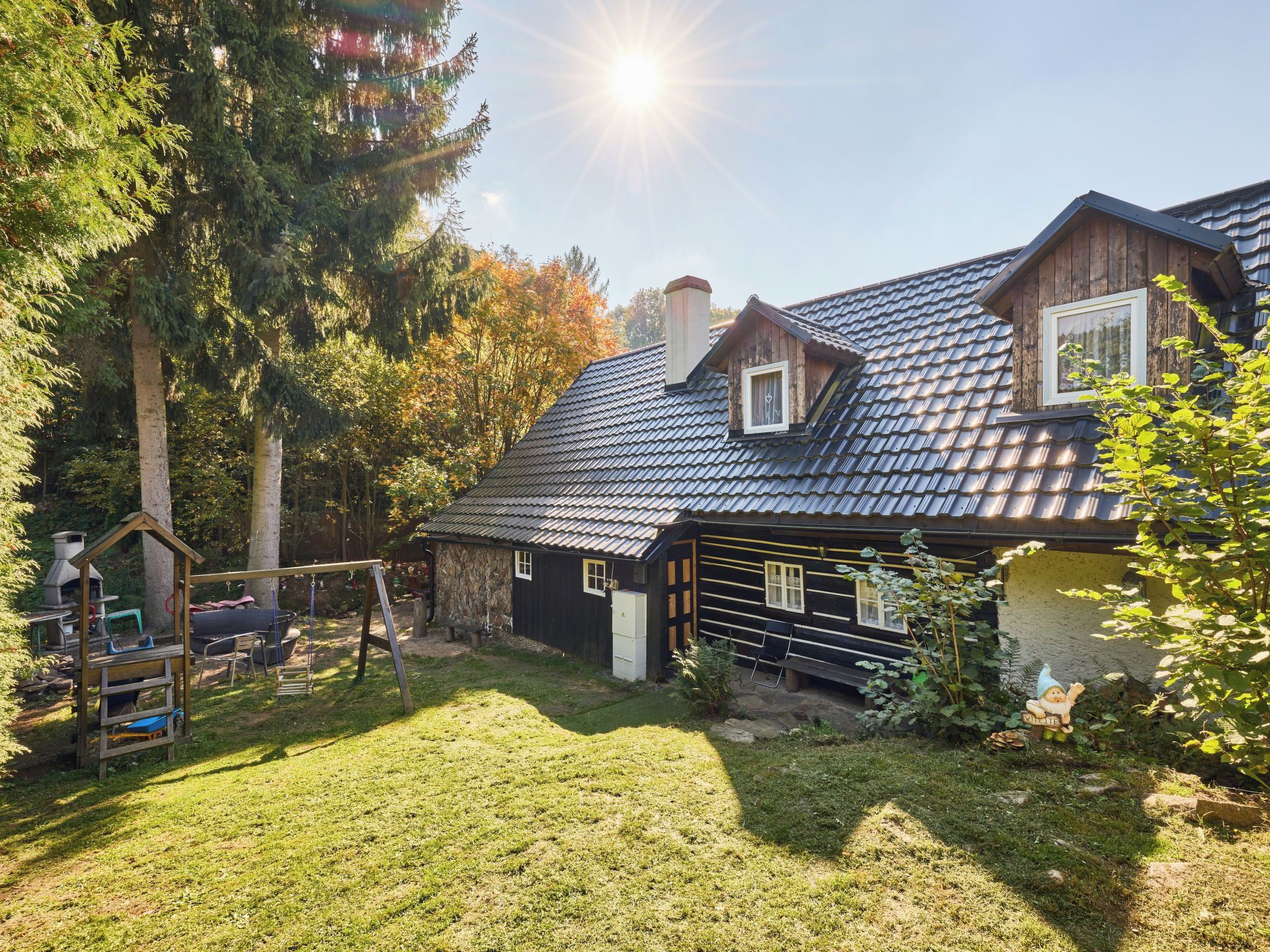 Une maison en bois avec un toit en pente et un petit abri attenant, entourée d’une cour verdoyante et d’arbres.