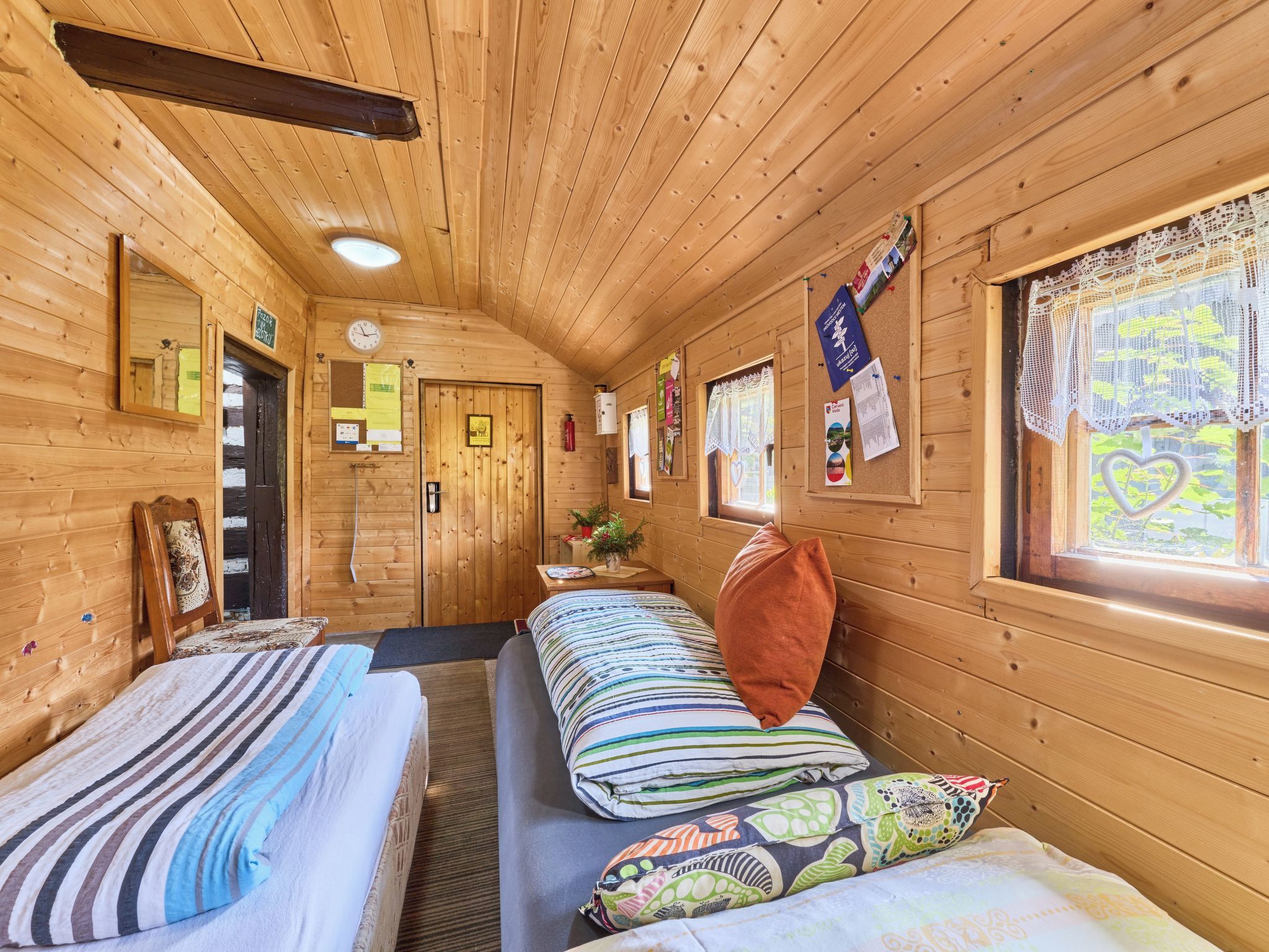 Een chalet met twee bedden, een houten plafond, een raam met kantgordijnen en een prikbord met verschillende papieren.
