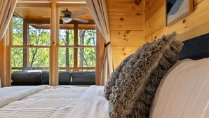 Een slaapkamer in een houten hutje, met een bed, een zacht kussen, een plafondventilator en uitzicht op bomen door grote ramen.