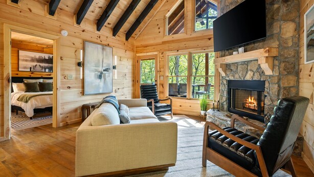 Een houten blokhutinterieur met een open haard, een bank, een leren fauteuil en een aan de muur bevestigde televisie.