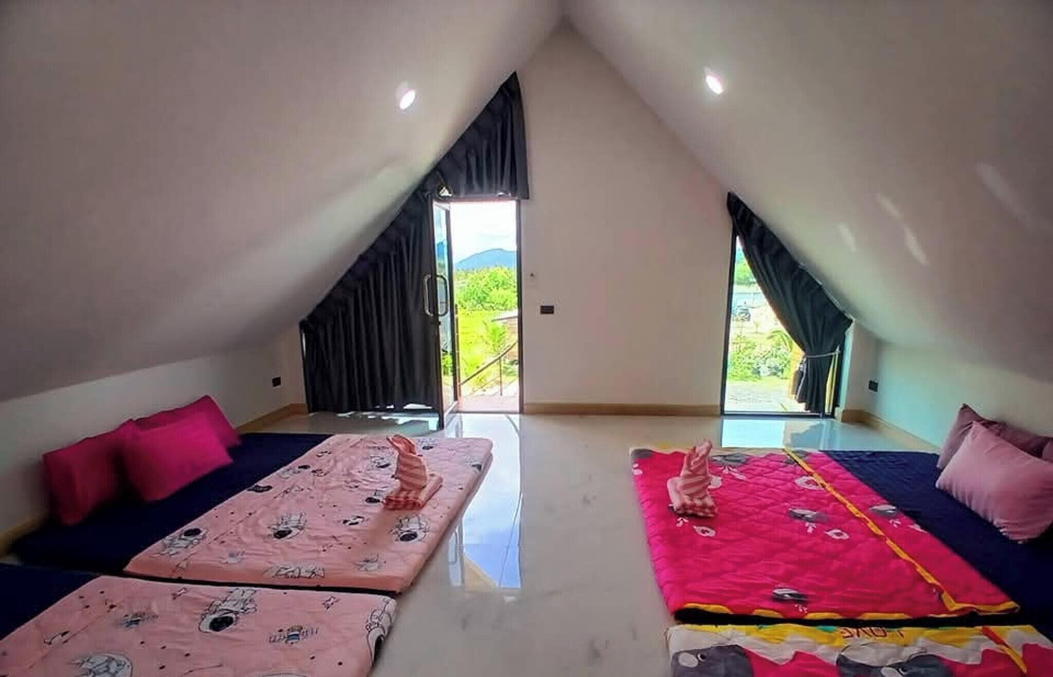Una stanza con soffitto alto, due letti – uno con copripiumino rosa e l’altro con copripiumino rosso – e un’ampia finestra che si affaccia sul verde circostante.