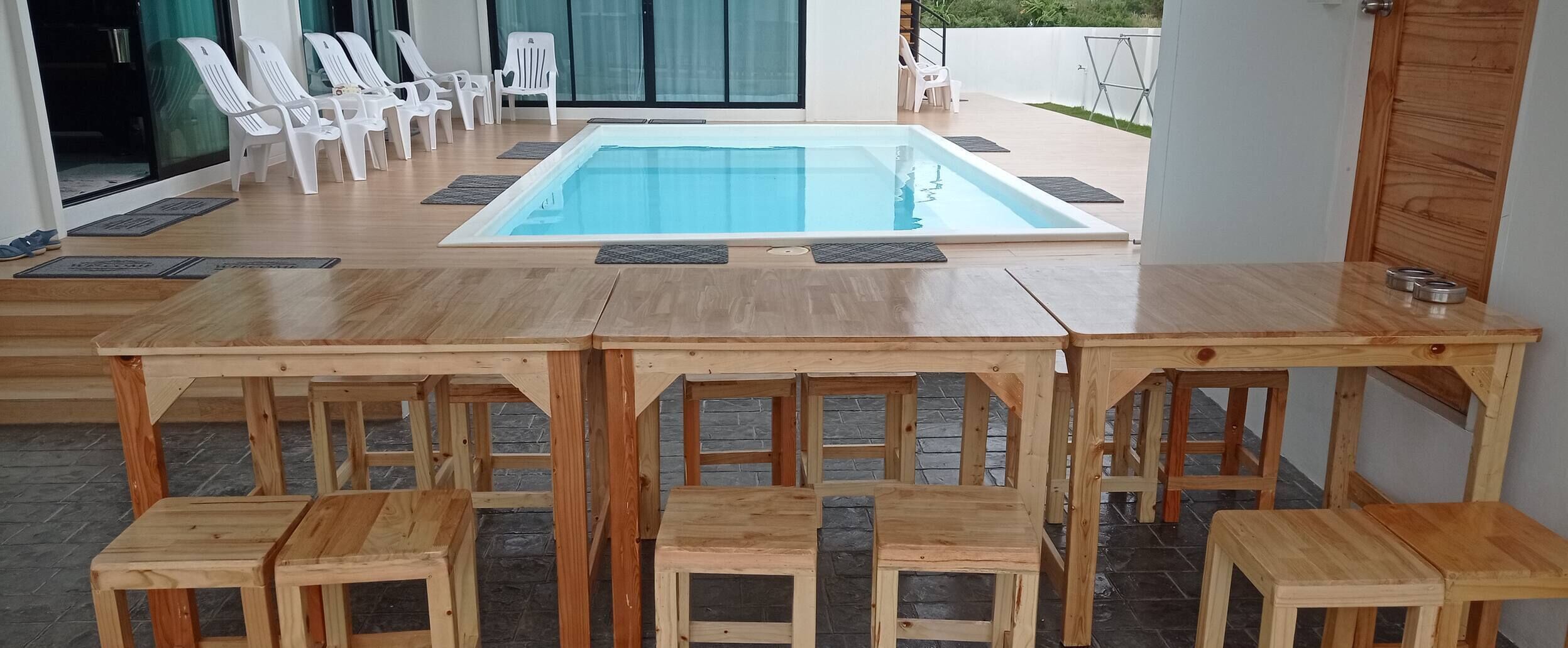 A poolside area with a wooden table and chairs, a clear blue pool, and a view of the outdoors.