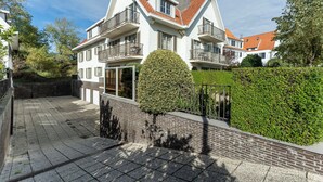 En tvåvånings bostadsbyggnad med ett tak täckt av takpannor, en balkong på andra våningen och en välskött häck på förgrunden.