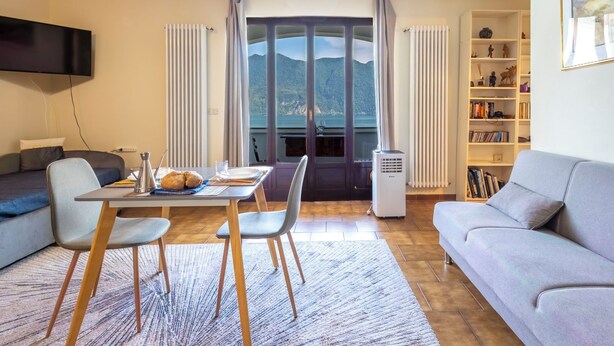 Ein Wohnzimmer mit Esstisch, Stühlen, einer Couch, einem Fernseher, einem Bücherregal und Bergblick durch ein Fenster.