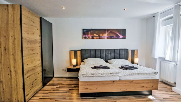 A modern bedroom with a wooden headboard, a bed with two pillows, a nightstand with a lamp, and a cityscape painting on the wall. - Fewo Römerweg by Interhome (Neustadt an der Weinstraße)