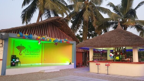 A small outdoor bar area with a thatched roof, a neon sign, and a seating area with a palm tree backdrop. - Elephant Garden Hotel and Resort (Kanchanrup)