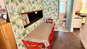 Un coin repas avec une table et des chaises, une étagère fixée au mur et une vue sur la cuisine.