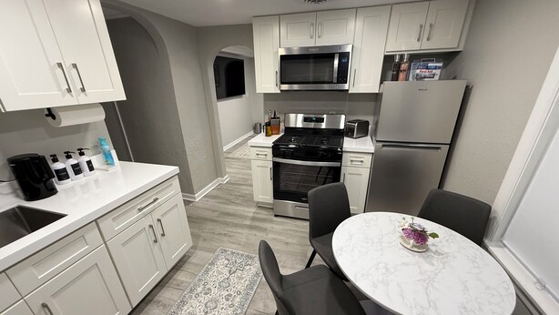 A modern kitchen with white cabinets, a built-in microwave, stove, and refrigerator. There is a dining area with a marble table and black chairs.