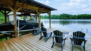 Een houten steiger met stoelen en een tafel, een overdekt gedeelte met een boot, en uitzicht op een meer.