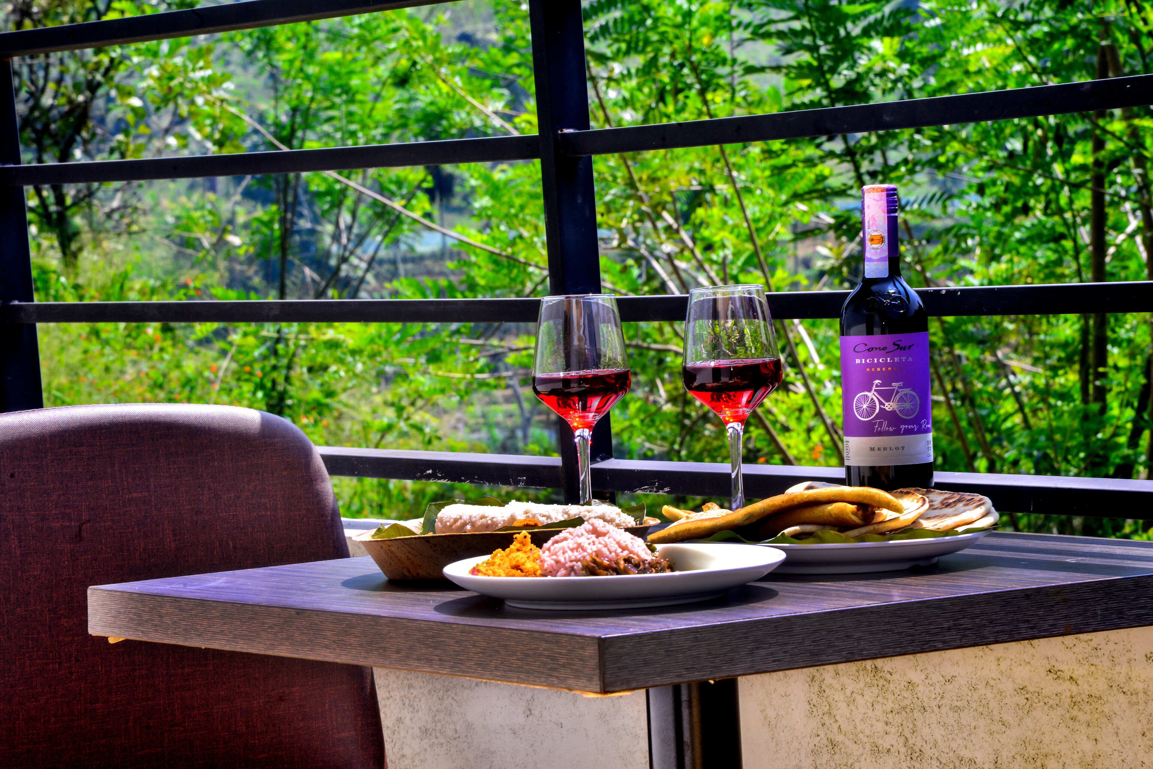 A table with plates of food, two wine glasses, and a bottle of wine, set against a backdrop of greenery.