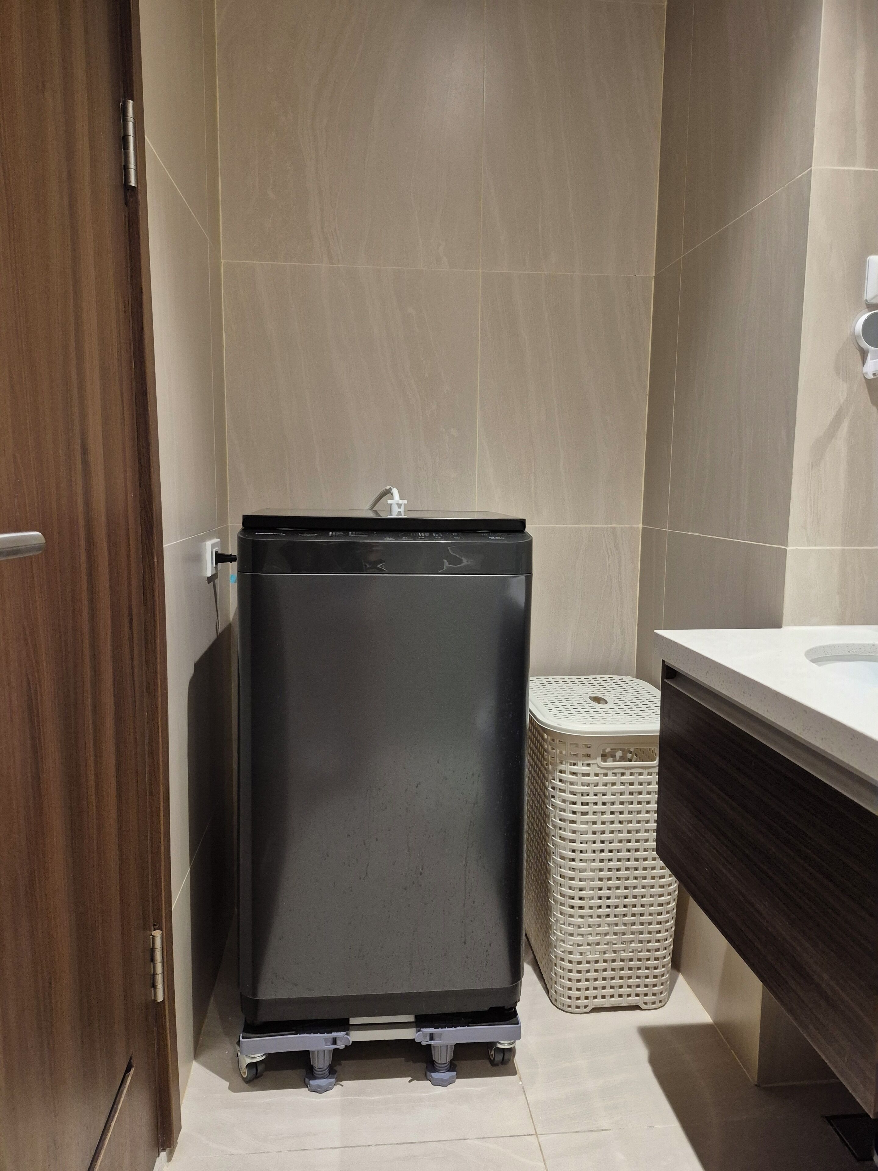 A compact bathroom with a small, dark-colored washing machine, a white laundry basket, and a modern sink.