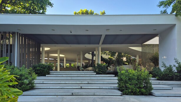 Un bâtiment moderne doté d’une entrée couverte, d’un escalier menant à l’entrée et entouré de verdure.