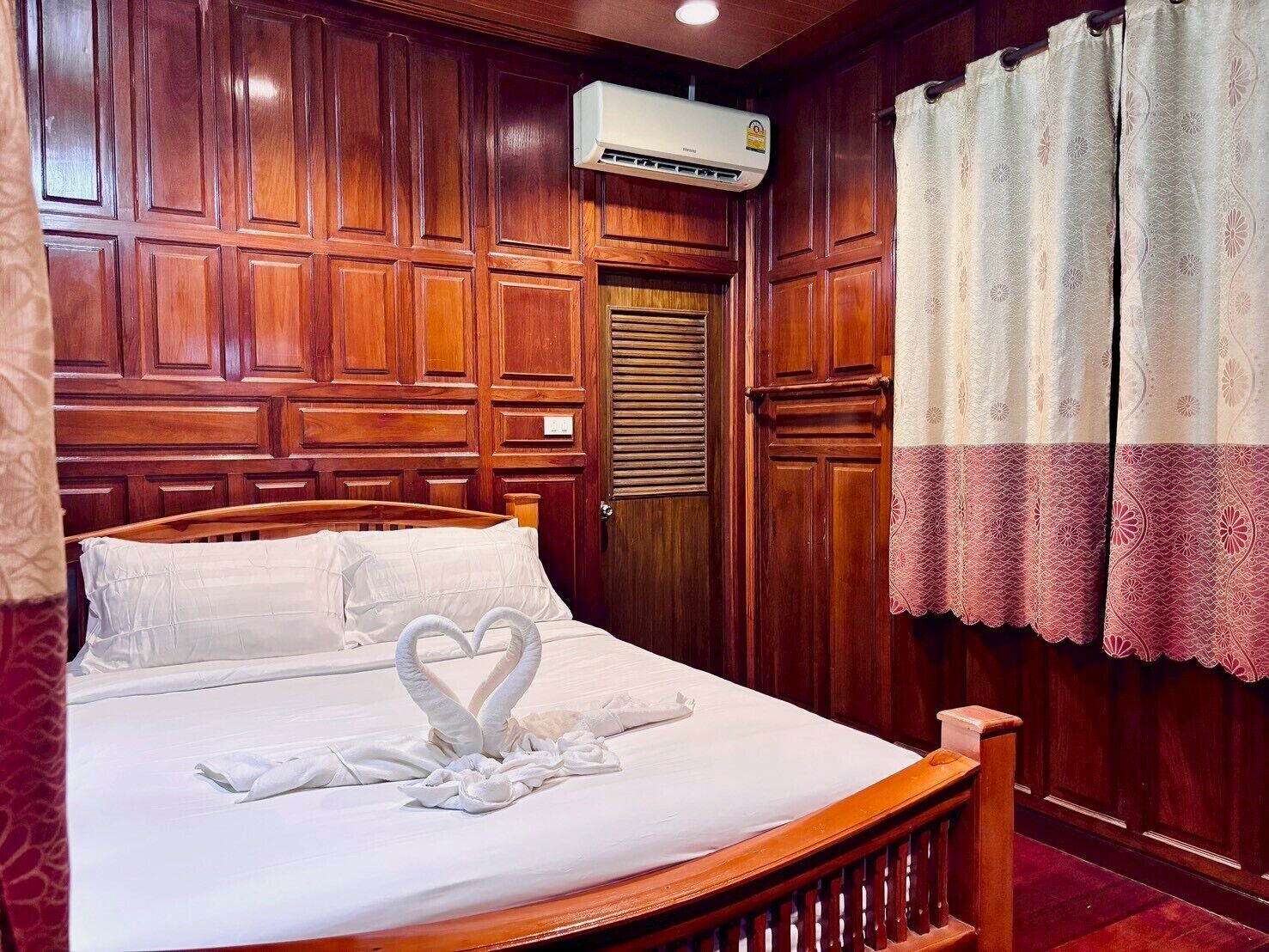 A hotel room with a wooden headboard, a bed with white linens, a heart-shaped towel arrangement, an air conditioning unit, and a window with patterned curtains.
