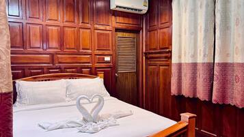 A hotel room with a wooden headboard, a bed with white linens, a heart-shaped towel arrangement, an air conditioning unit, and a window with patterned curtains.