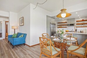 A modern living room with a blue sofa, wicker chairs, a dining table set for four, and a kitchen area with a refrigerator and microwave. - Kapalua Ridge | Ocean View 2 BR, Sleeps 8 | Car Incl. w/ 6+ Nights | KRV ML-3942 by KBM (Lahaina)