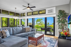 A modern living room with a gray sofa, a wooden coffee table, and a ceiling fan. Large windows offer a view of greenery and a distant landscape. - Kapalua Ridge | Ocean View 2 BR, Sleeps 8 | Car Incl. w/ 6+ Nights | KRV ML-3942 by KBM (Lahaina)