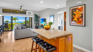 A modern kitchen with a central island, bar stools, a dining area with a view, and a living space with a sofa. - Kapalua Ridge | Ocean View 3 BR, Sleeps 12 | Car Incl. w/ 6+ Nights | KRV ML-3963 by KBM (Lahaina)