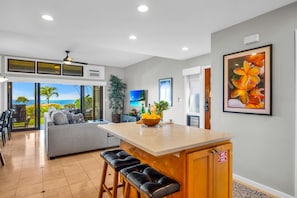 A modern kitchen with a central island, bar stools, a dining area with a view, and a living space with a sofa. - Kapalua Ridge | Ocean View 3 BR, Sleeps 12 | Car Incl. w/ 6+ Nights | KRV ML-3963 by KBM (Lahaina)