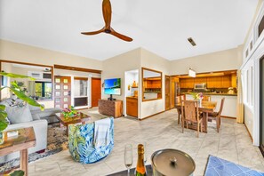 A spacious living area with a sofa, coffee table, and a TV. There is a dining area with a table and chairs. The kitchen has wooden cabinets and a microwave. - Kapalua Ridge | Ocean View 3 BR, Sleeps 12 | Car Incl. w/ 6+ Nights | KRV ML-3963 by KBM (Lahaina)