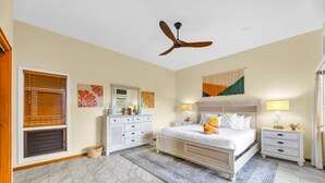 A bedroom with a bed, two bedside tables, a ceiling fan, a large window with blinds, and a decorative wall hanging. - Kapalua Ridge | Ocean View 3 BR, Sleeps 12 | Car Incl. w/ 6+ Nights | KRV ML-3963 by KBM (Lahaina)