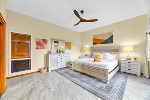 A bedroom with a bed, two bedside tables, a ceiling fan, a large window with blinds, and a decorative wall hanging. - Kapalua Ridge | Ocean View 3 BR, Sleeps 12 | Car Incl. w/ 6+ Nights | KRV ML-3963 by KBM (Lahaina)
