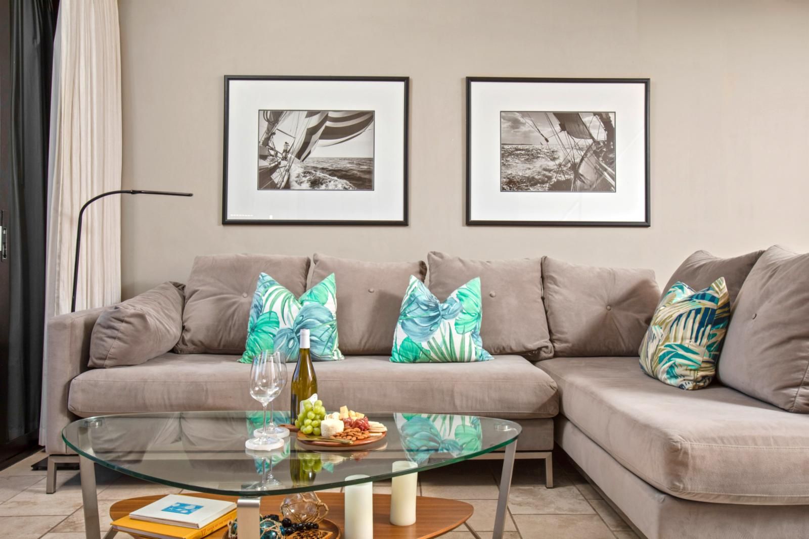 A living room with a beige sofa, two framed black and white nautical paintings, a glass coffee table with a bottle of wine, grapes, and cheese, and tropical patterned pillows.