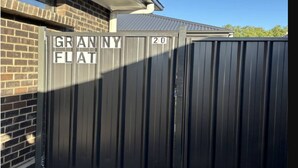 Una reja metálica con los letreros 'GRANNY FLAT' y '20', un edificio de ladrillo y un cielo despejado.