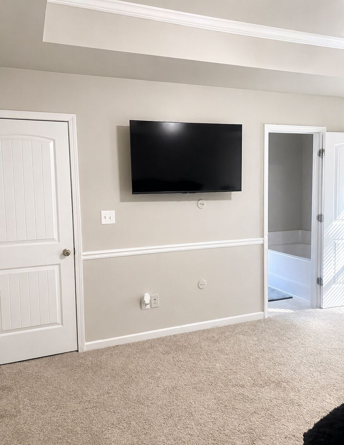A room with a flat-screen TV mounted on the wall, a white door, and a carpeted floor.