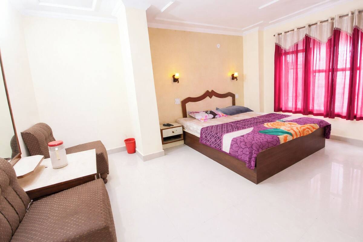 Habitación de hotel con cama, una mesita, una silla, una mesita de noche y un ventanal con cortinas.