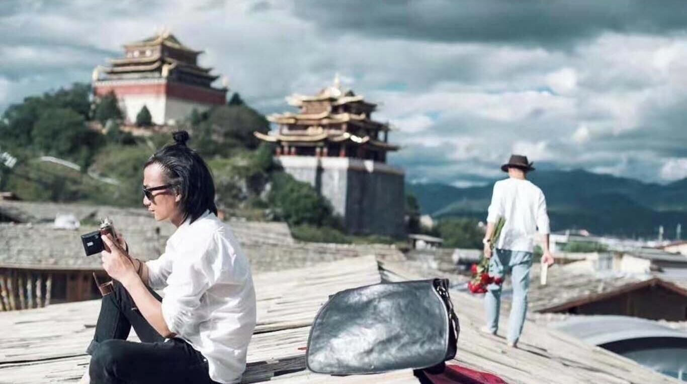 Una persona sentada en una terraza de madera, con una cámara, observando edificios tradicionales y un paisaje montañoso.
