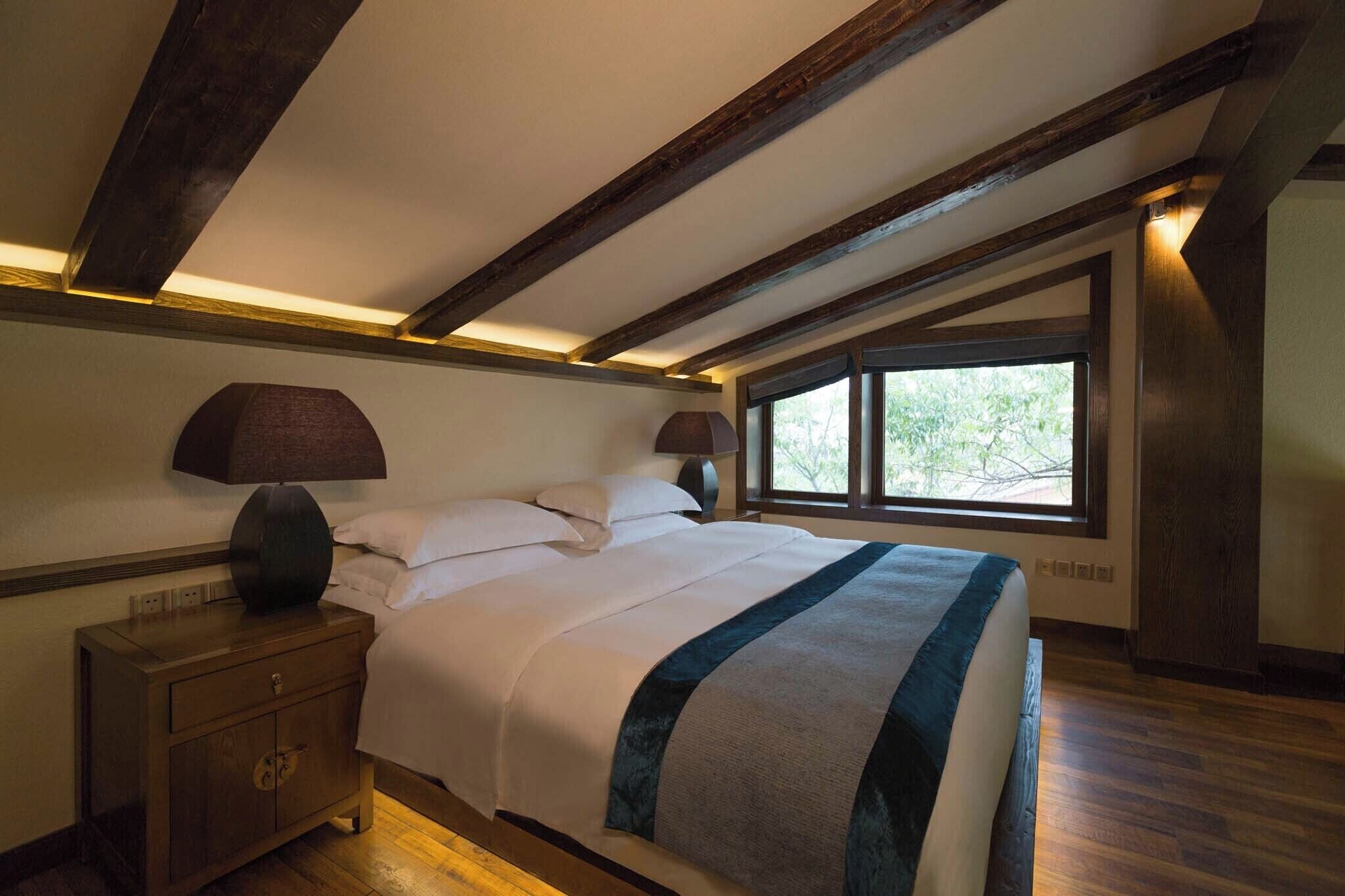 A bedroom with a large bed, wooden beams, a nightstand, a lamp, and a window with a view.