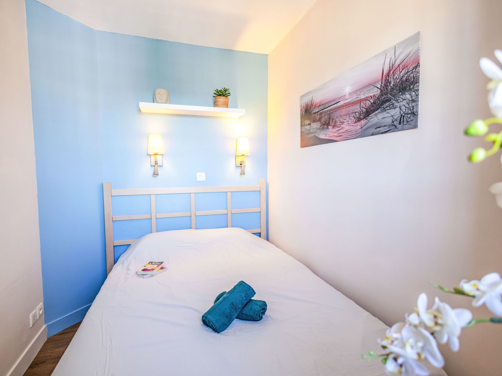 Un dormitorio con cama, cabecera, repisa con planta, luces fijadas a la pared y un cuadro.