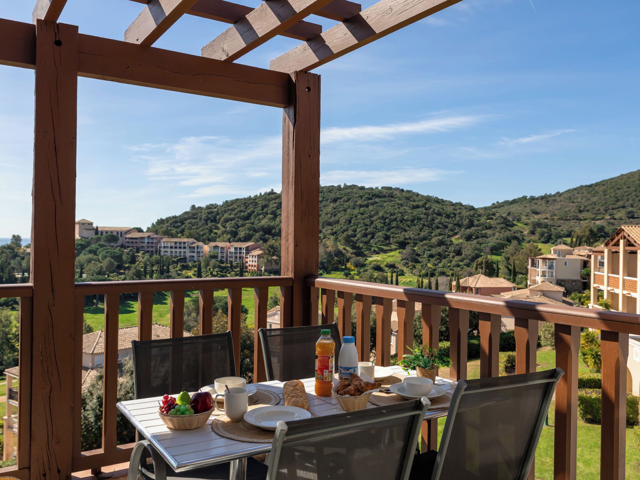 Varanda de madeira com mesa posta para café da manhã, com vista para uma colina e edifícios.