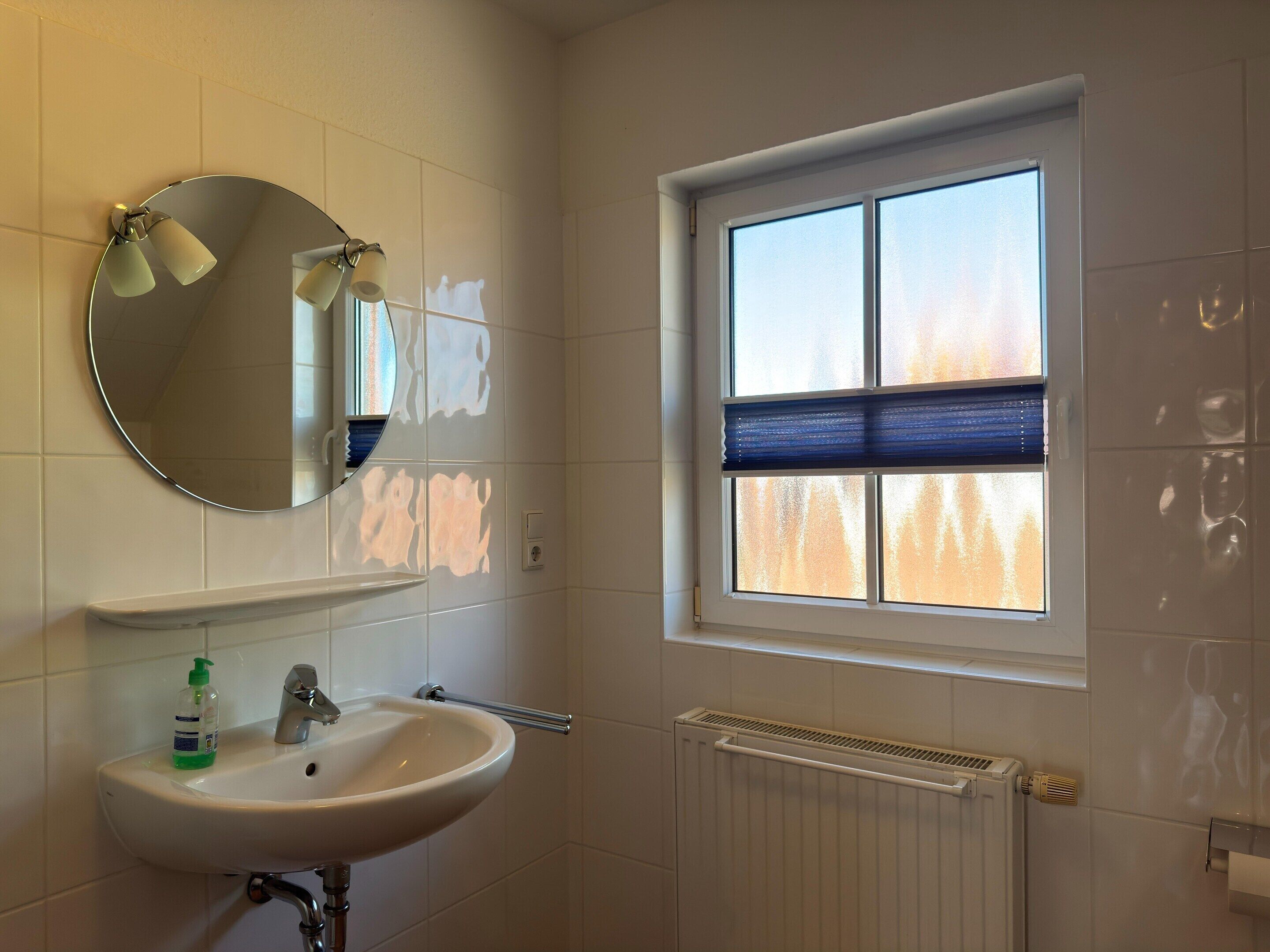Et badeværelse med en hvid vask, et spejl, et vindue med skodder og en radiator.